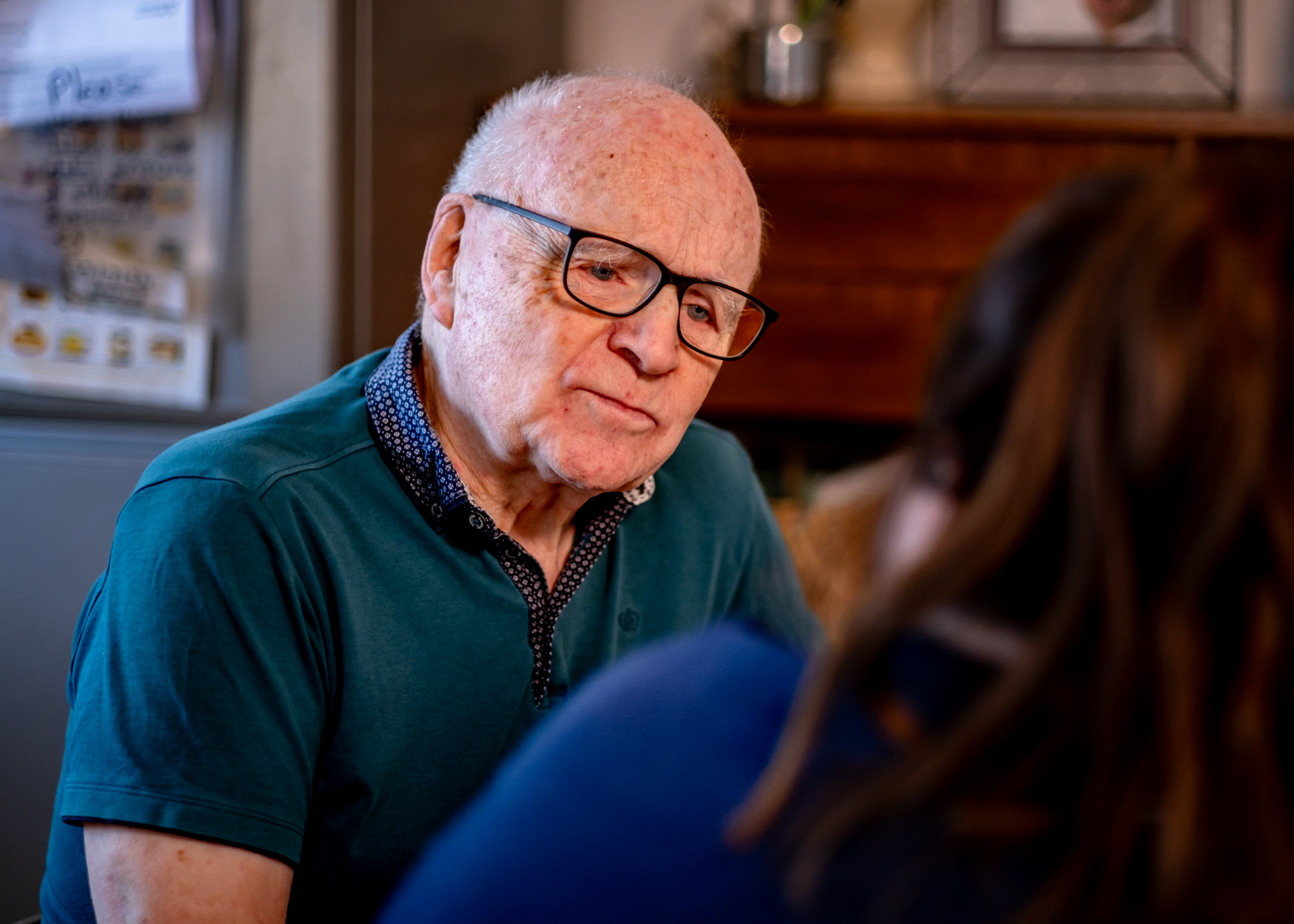 An older man wearing glasses and a green polo shirt in deep conversation with an out of focus carer from Apollo Care Liverpool