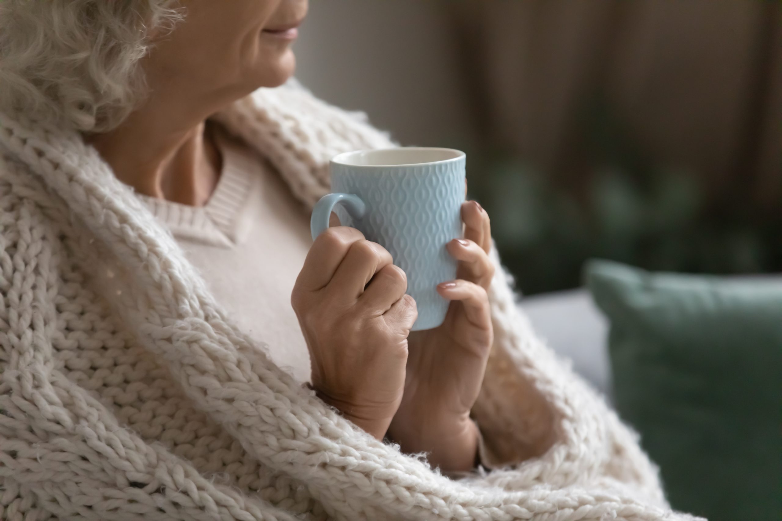 An older lady keeps herself warm with a thick cardigan and a mug of hot drink.
