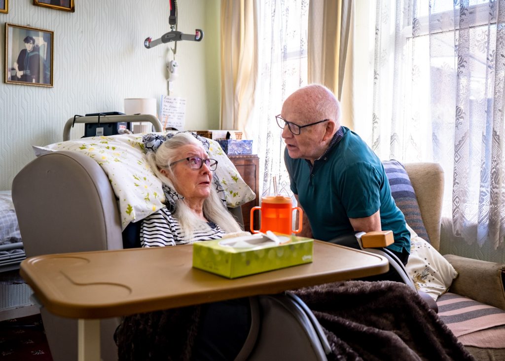 An older man in a green polo shirt, tenderly talking to his wife who's sitting in an armchair