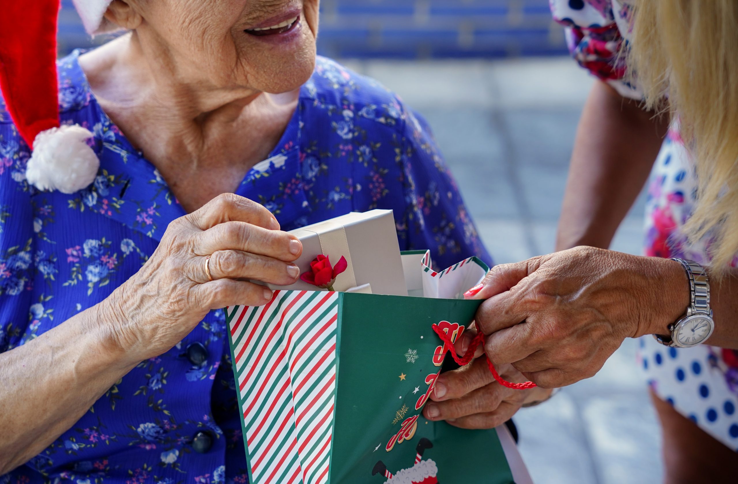 An older lady wearing a blur top and santa hat receiving a small Christmas gift from an out of shot friend.