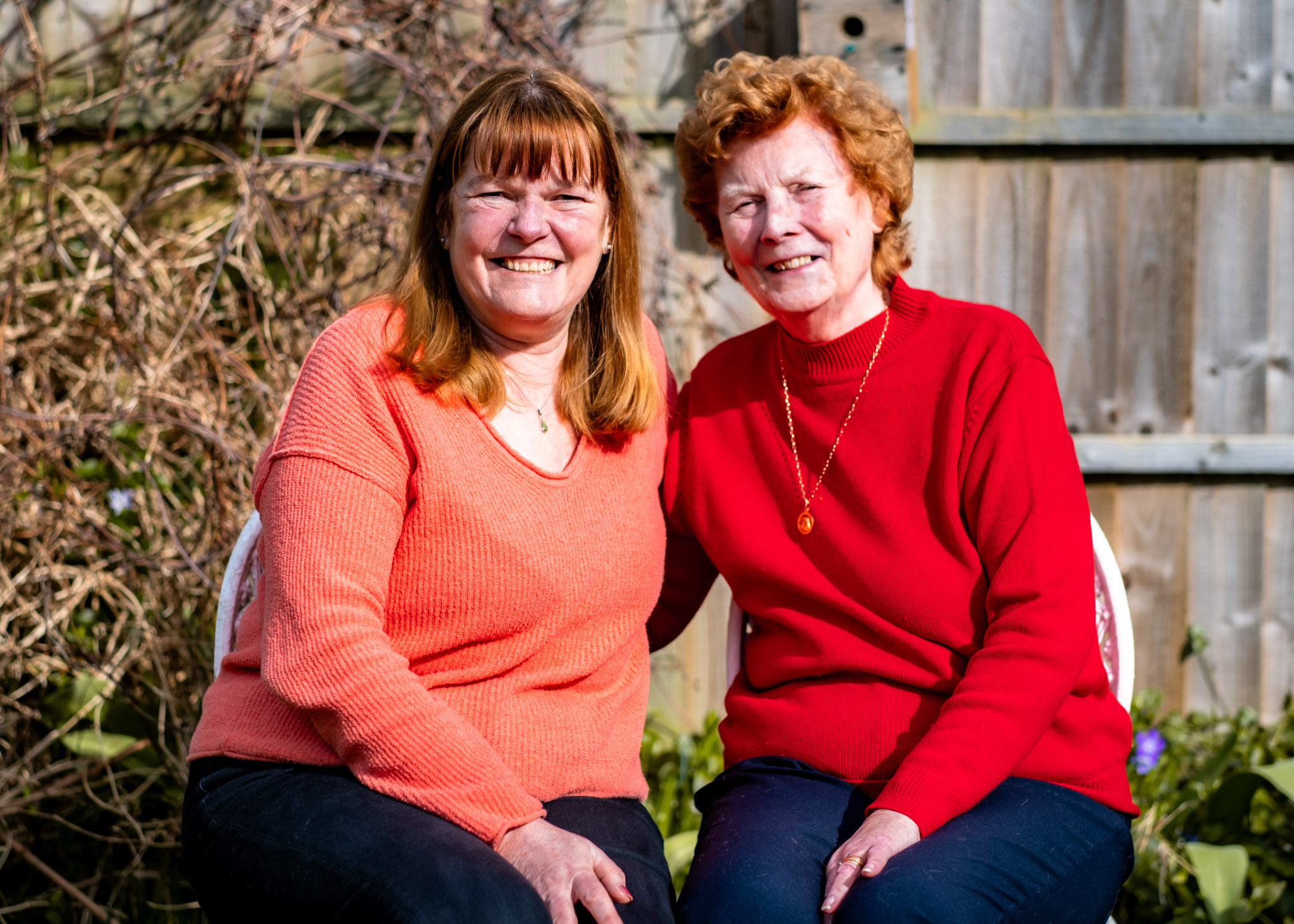 A middle-aged daughter sitting with in the garden with her mother, smiling into the camera.